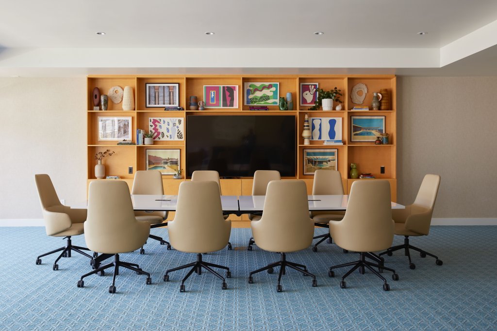 Group Incentive A modern conference room with a sleek, white table surrounded by ten beige office chairs on a blue patterned carpet, featuring a large built-in wooden bookshelf filled with colorful artwork and decorative items against a white wall in Santa Clara, California.