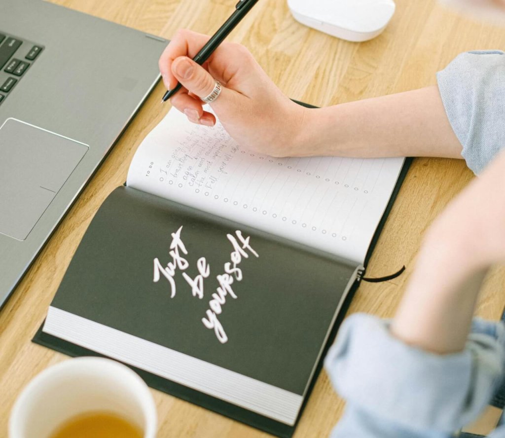 A woman writes notes in an open notebook with 'Just Be Yourself' on the cover in a cozy home office setting.