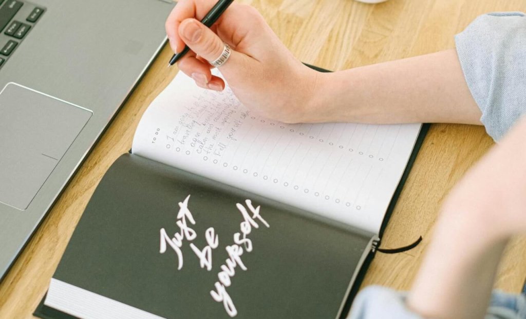 A woman writes notes in an open notebook with 'Just Be Yourself' on the cover in a cozy home office setting.