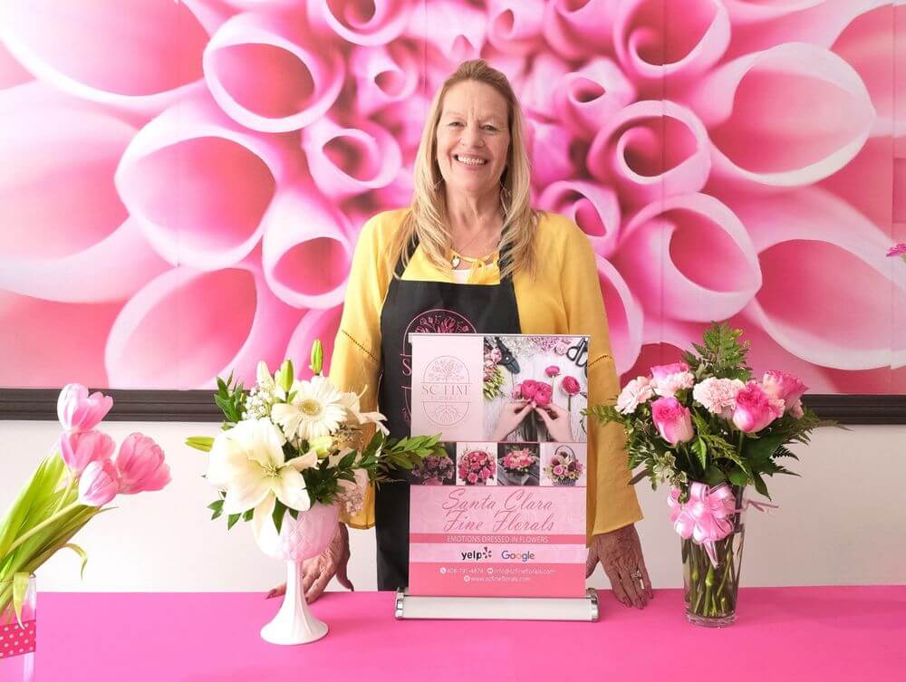 A woman in a yellow shirt and black apron poses with flowers in front of a pink floral backdrop in Santa Clara, California.