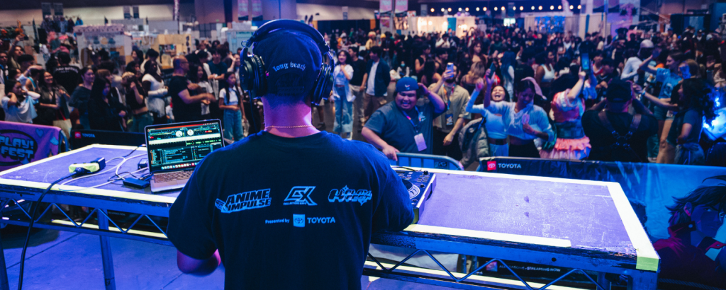 A DJ at a bustling anime festival, wearing headphones and a black t-shirt, operating a DJ booth with a laptop and DJ equipment in Santa Clara, California.