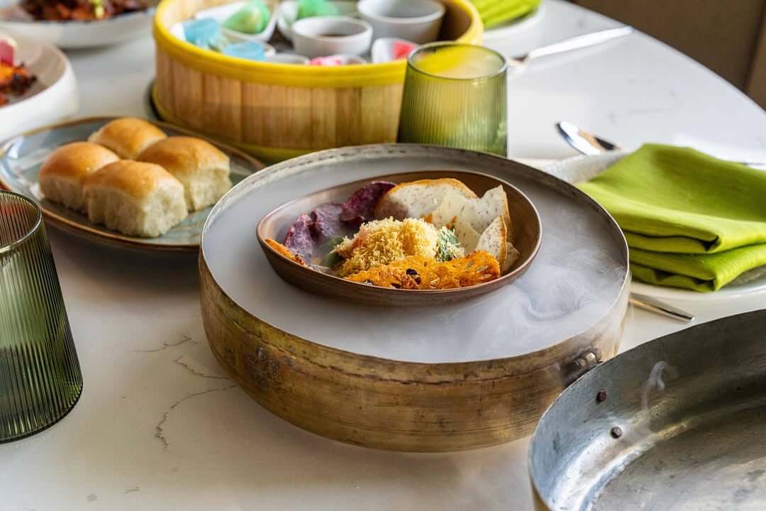 A variety of colorful dishes, including steamed buns, a bowl of food in a copper vessel with smoke rising from it, and a yellow bamboo steamer filled with small bowls, are arranged on a white table with green napkins and glasses in Santa Clara, California.