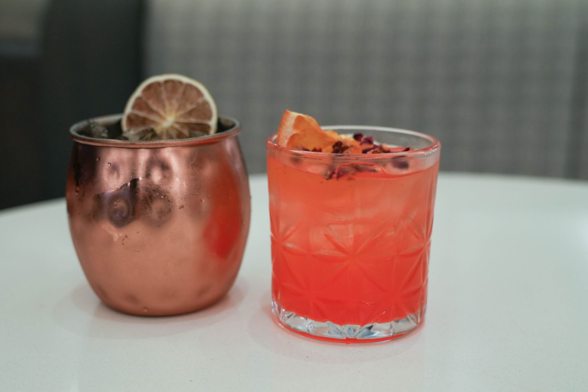A copper Moscow Mule mug filled with a dark liquid and garnished with a dried lime slice, alongside a tall glass filled with a vibrant red cocktail and topped with an orange slice and rose petals in Santa Clara, California.