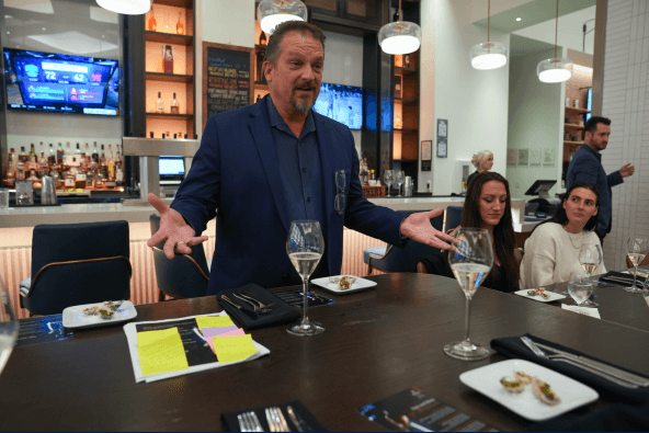 A man in a blue suit gestures while speaking to a group seated at a table in a modern restaurant setting in Santa Clara, California.