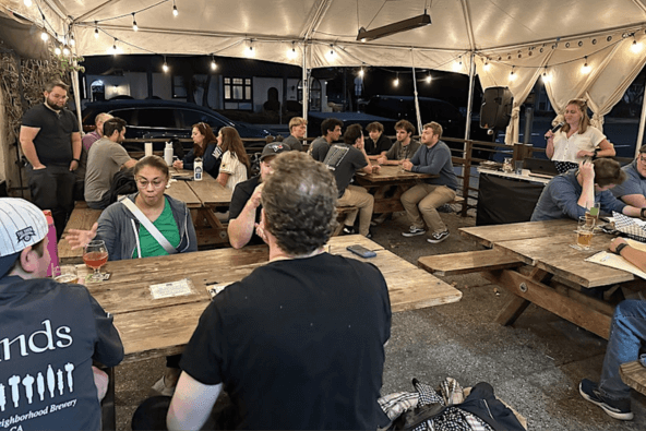A group of people is gathered under a large tent, seated at wooden picnic tables, enjoying drinks and conversation in Santa Clara, California.