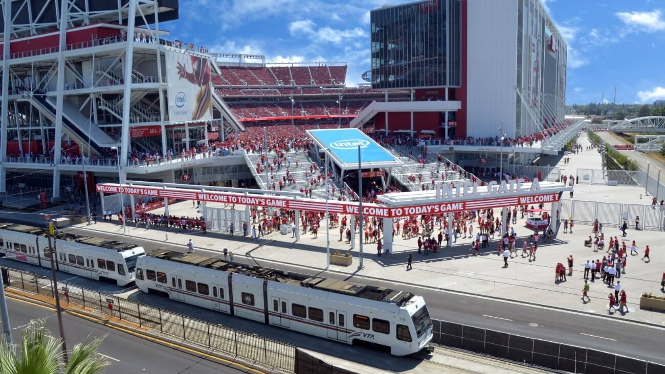 Crowded entrance to a stadium, with fans gathering and a train passing by in Santa Clara, California.