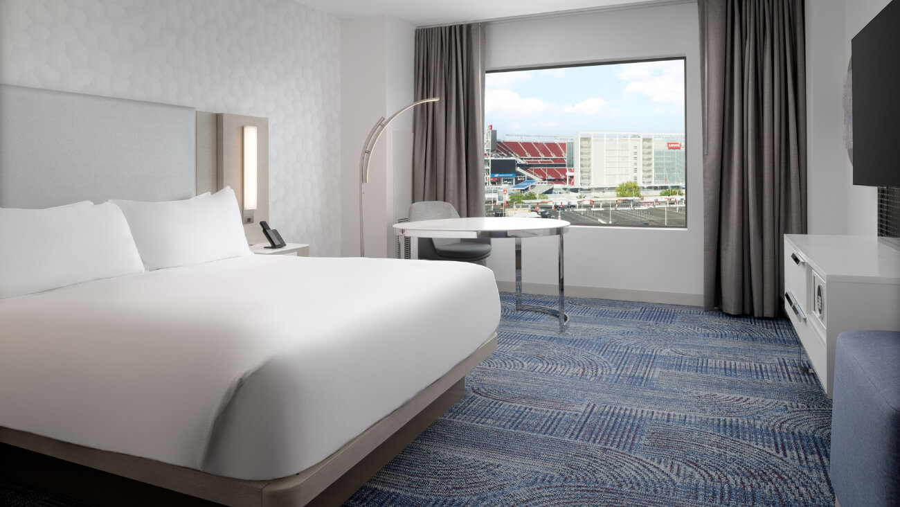 A modern hotel room with a large bed, a desk, and a window overlooking a cityscape in Santa Clara, California.