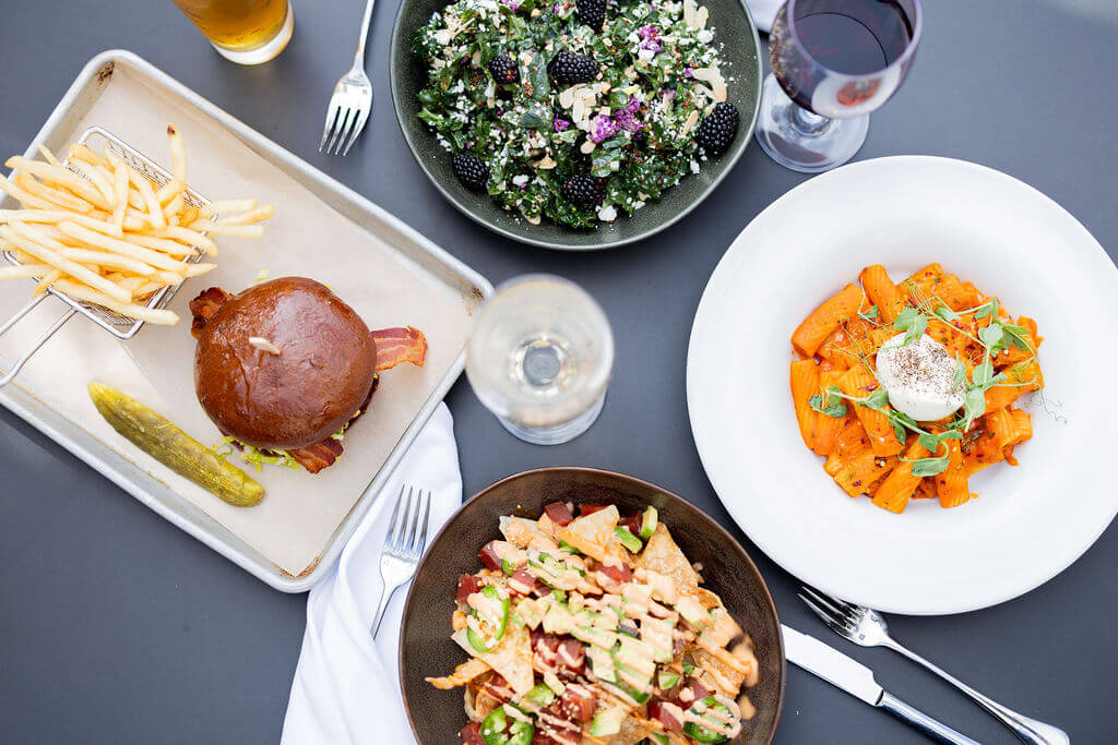 A variety of dishes including a burger with fries, a kale salad, pasta with red sauce, and nachos with avocado and jalapeños, accompanied by wine and beer in Santa Clara, California.