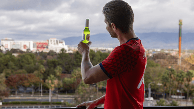 A man in a red sports jersey holds a beer bottle, gazing at a cityscape with mountains in the distance in Santa Clara, California.