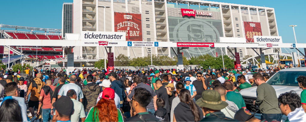 A massive crowd of people, many wearing green and red, gather outside a large stadium with a prominent Levi's sign, under a clear blue sky in Santa Clara, California.