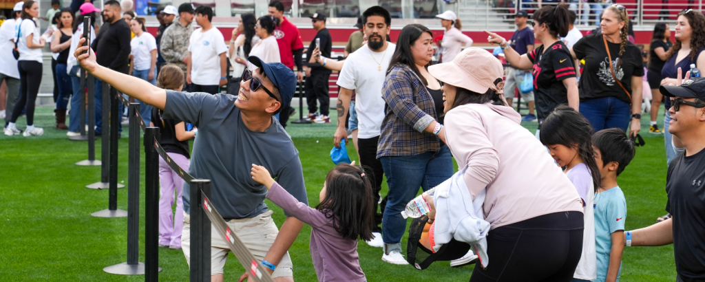 A lively crowd of people, including families and children, are gathered on a grassy field, taking selfies and enjoying a sunny day at an outdoor event in Santa Clara, California.