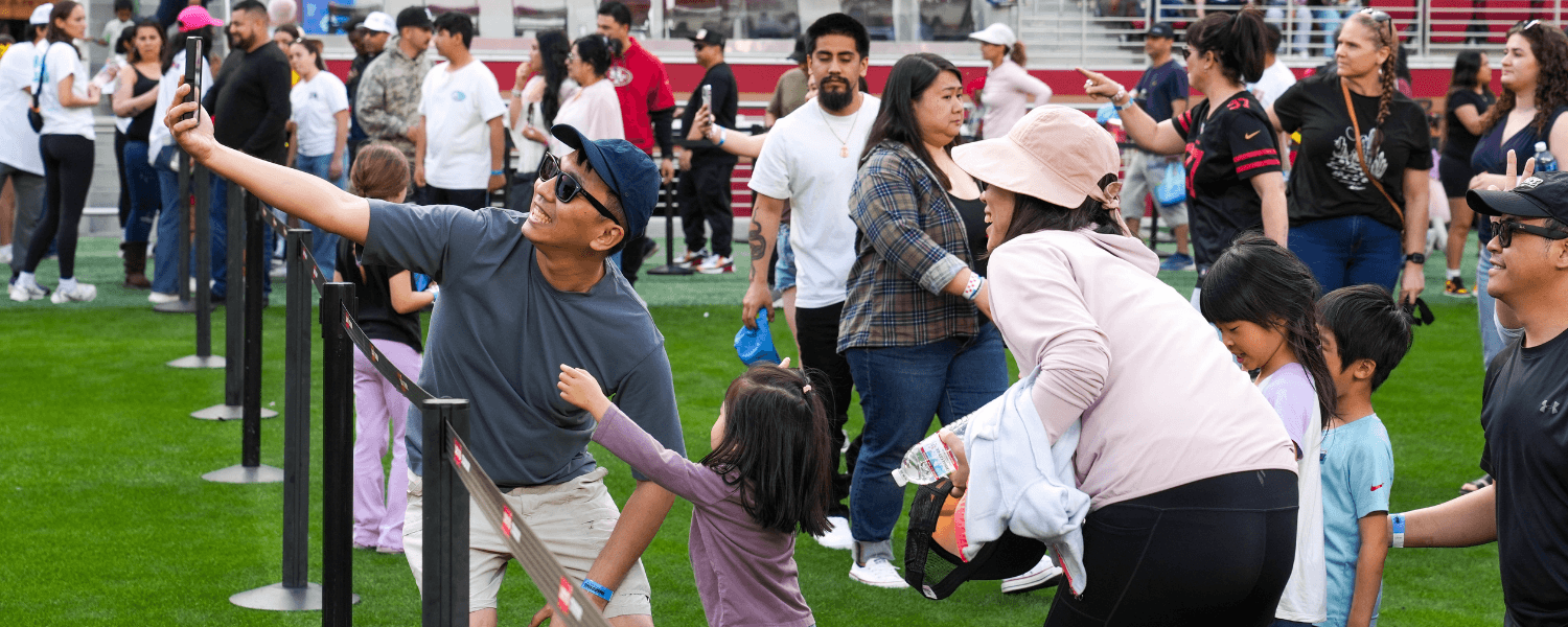 May Events in Santa Clara 2026 A lively crowd of people, including families and children, are gathered on a grassy field, taking selfies and enjoying a sunny day at an outdoor event in Santa Clara, California.