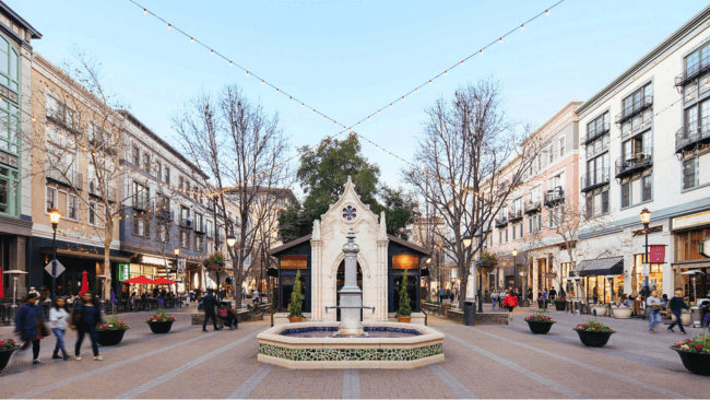 A bustling outdoor shopping center with a central fountain, surrounded by colorful buildings and filled with people in Santa Clara, California.