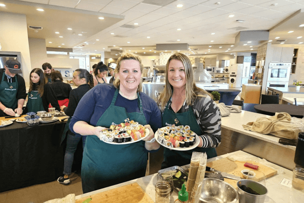 Two women in green aprons proudly display colorful sushi platters in a bustling kitchen in Santa Clara, California.