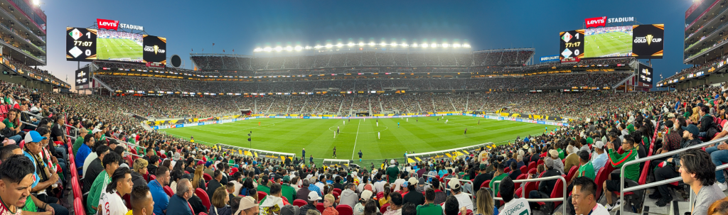 A panoramic view of a packed stadium during a soccer match, with bright lights illuminating the field and enthusiastic fans filling the stands in Santa Clara, California.