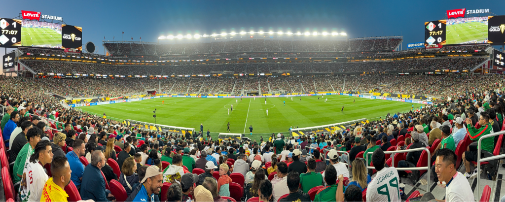 A panoramic view of a packed stadium during a soccer match, with bright lights illuminating the field and enthusiastic fans filling the stands in Santa Clara, California.