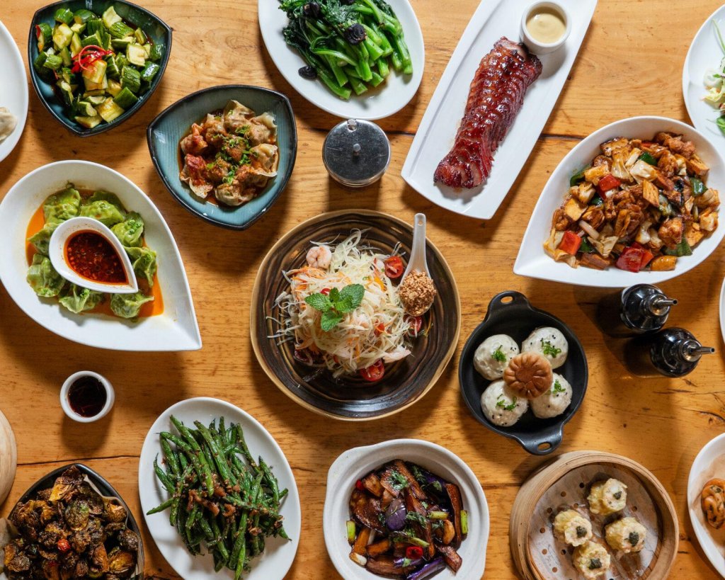 A variety of colorful and appetizing Asian dishes, including stir-fries, soups, dumplings, and grilled meats, are spread across a wooden table in Santa Clara, California.