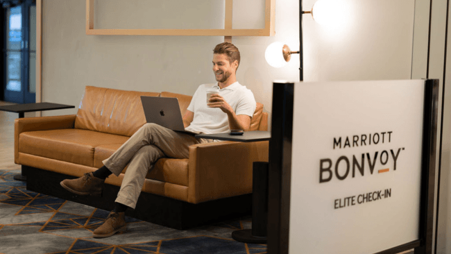 A man relaxes on a brown leather couch with a laptop and coffee, near a Marriott Bonvoy Elite Check-In sign in Santa Clara, California.