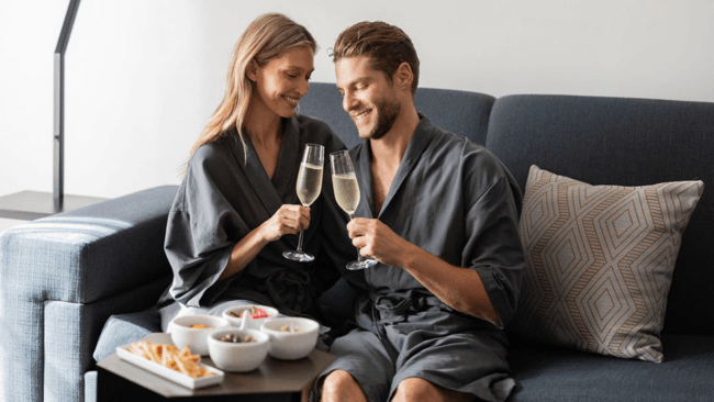 A couple in matching gray robes enjoys a romantic moment on a couch, toasting with champagne flutes in Santa Clara, California.
