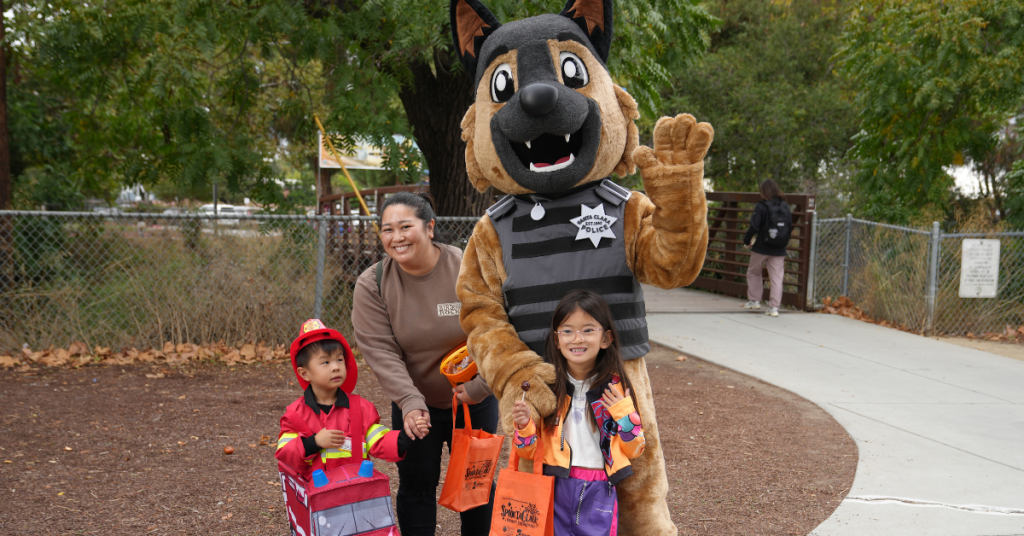 Children's SpooktaClara Halloween Party A woman and two children in costumes pose with a police dog mascot outdoors in Santa Clara, California.